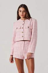 A woman is wearing a pink, textured, tailored two-piece outfit consisting of a cropped jacket with structured shoulders and decorative buttons, and matching high-waisted shorts. The fabric has a woven pattern with shades of pink and white.